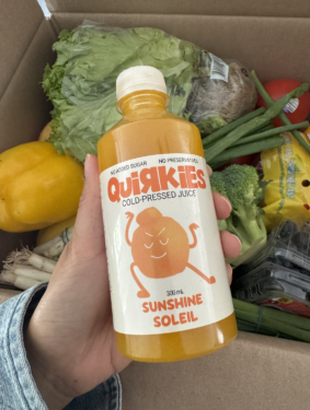 Hand holding a bottle of orange Odd Bunch Quirkies Organic Cold Pressed Juice with groceries in the background